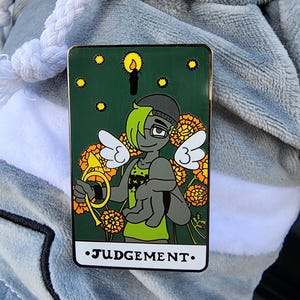 May include: A rectangular enamel pin featuring a cartoon character with angel wings, glasses, and a green and gray color scheme. The pin has a dark green background with yellow flowers and stars, and the word "JUDGEMENT" at the bottom.