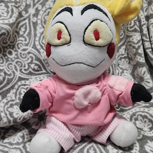 Pode incluir: Uma boneca de pelúcia com um rosto branco, cabelo amarelo e detalhes vermelhos ao redor dos olhos. Ela usa uma camisa rosa e calças listradas. A boneca tem mãos pretas e pés brancos. A boneca está sentada em um cobertor estampado.