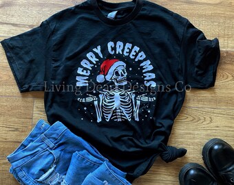 May include: Black t-shirt with a white skeleton graphic wearing a Santa hat. The text "MERRY CREEPMAS" is arched above the skeleton. The shirt is paired with blue jeans and black boots.