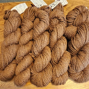 May include: Several skeins of brown yarn are displayed, neatly arranged on a wooden surface. The yarn is thick and textured, with tags attached. The color is a deep, earthy brown, suitable for various knitting projects.