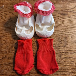 May include: A pair of white doll shoes with red lace trim at the top and a textured floral pattern on the toe, accompanied by a pair of solid red knit socks. The shoes feature a white fabric upper and a molded white sole with a rounded toe design.
