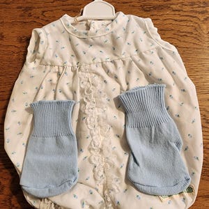 May include: A white baby romper with a blue floral pattern and lace trim, paired with two light blue knitted mittens. The romper is hanging on a white plastic hanger.