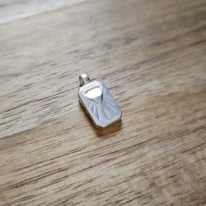 Op de afbeelding: Een zilveren hanger met een geometrisch ontwerp. De rechthoekige hanger heeft een driehoek en lijnen die in het metaal zijn geëtst. De hanger heeft een kleine lus aan de bovenkant voor een ketting. De hanger wordt gepresenteerd op een houten oppervlak.