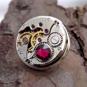 May include: A silver ring showcasing the detailed mechanism of a watch, with visible golden gears and a bright red gemstone. The ring's design includes small, intricate parts, offering a unique, industrial look. The ring is set against a textured, brown surface.