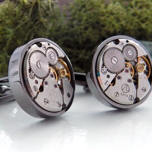 May include: A pair of silver cufflinks with exposed watch gears. The round faces of the cufflinks display intricate clockwork mechanisms with silver and gold-colored components. The cufflinks are set against a white surface with a green moss background.