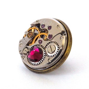 May include: A circular, antique-style watch face with a pink gemstone in the centre. The watch face is surrounded by a brass-coloured frame and features intricate gears and markings.