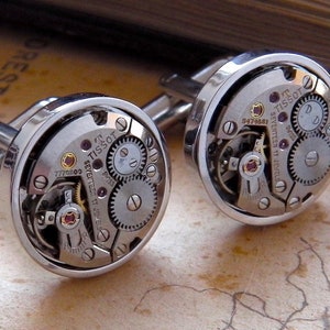 May include: Silver cufflinks showcasing the intricate internal mechanisms of a watch. The design reveals gears, jewels, and the Tissot brand. These round cufflinks offer a distinctive look, ideal for formal wear. The watch mechanisms are visible.