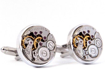 Puede incluir: Un par de gemelos de plata con un diseño circular que muestra un mecanismo de reloj visible con engranajes dorados y joyas de color púrpura.