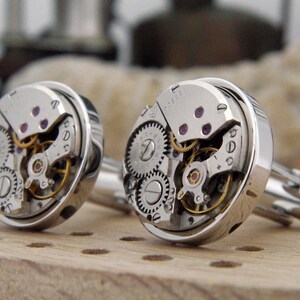 Puede incluir: Un par de gemelos de plata con mecanismos de reloj expuestos. Los gemelos tienen un dise&ntilde;o circular, con una carcasa de plata y una cara transparente que muestra el funcionamiento interno de un reloj.