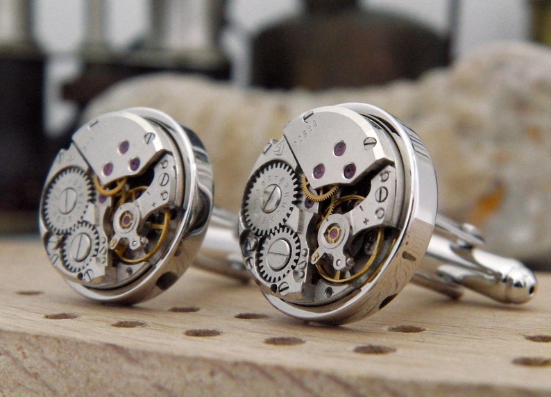 Puede incluir: Un par de gemelos de plata con mecanismos de reloj expuestos. Los gemelos tienen un dise&ntilde;o circular, con una carcasa de plata y una cara transparente que muestra el funcionamiento interno de un reloj.