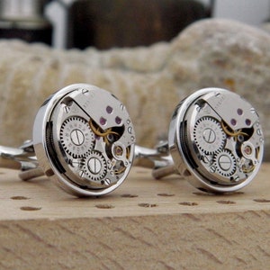 Puede incluir: Un par de gemelos de plata con un dise&ntilde;o circular. Cada gemelo presenta un movimiento de reloj visible con engranajes y un borde plateado.