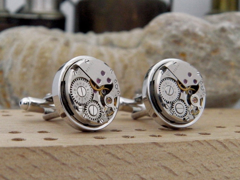 Puede incluir: Un par de gemelos de plata con un dise&ntilde;o circular. Cada gemelo presenta un movimiento de reloj visible con engranajes y un borde plateado.
