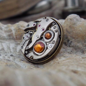 May include: A steampunk-style ring featuring the intricate inner workings of a watch. The ring has a bronze-colored bezel, with visible gears, jewels, and an orange-colored gemstone. The design showcases the mechanical beauty of timekeeping.