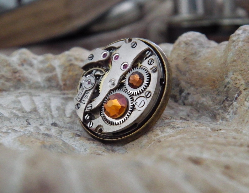 May include: A steampunk-style ring featuring the intricate inner workings of a watch. The ring has a bronze-colored bezel, with visible gears, jewels, and an orange-colored gemstone. The design showcases the mechanical beauty of timekeeping.