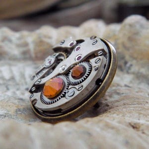 May include: A steampunk-style ring featuring the intricate inner workings of a watch. The ring has a silver-colored metal base with visible gears, and is accented with orange and red gemstones. The ring is set on a textured, light-colored surface.