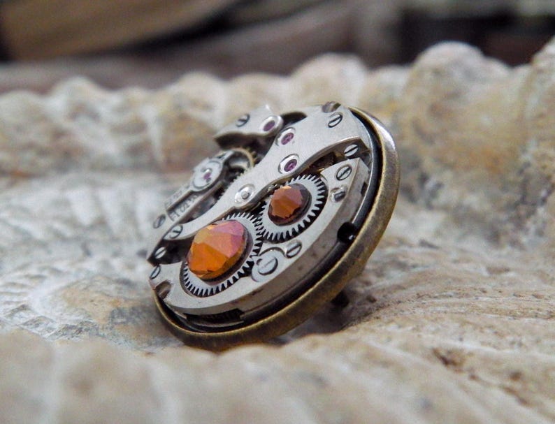 May include: A steampunk-style ring featuring the intricate inner workings of a watch. The ring has a silver-colored metal base with visible gears, and is accented with orange and red gemstones. The ring is set on a textured, light-colored surface.