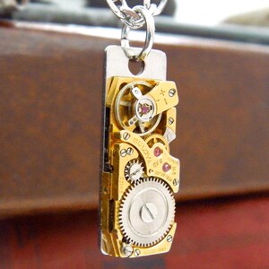 May include: A rectangular pendant featuring the intricate inner workings of a gold-toned watch. The pendant showcases gears, jewels, and other mechanical components, suspended from a silver chain. The design highlights the beauty of watchmaking.
