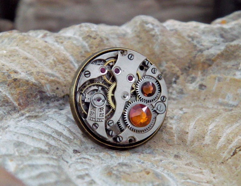 May include: A round, steampunk-style ring featuring the intricate inner workings of a watch. The ring has a silver-colored face with gears, jewels, and small screws. The setting is a bronze color. The ring has a unique, industrial aesthetic.