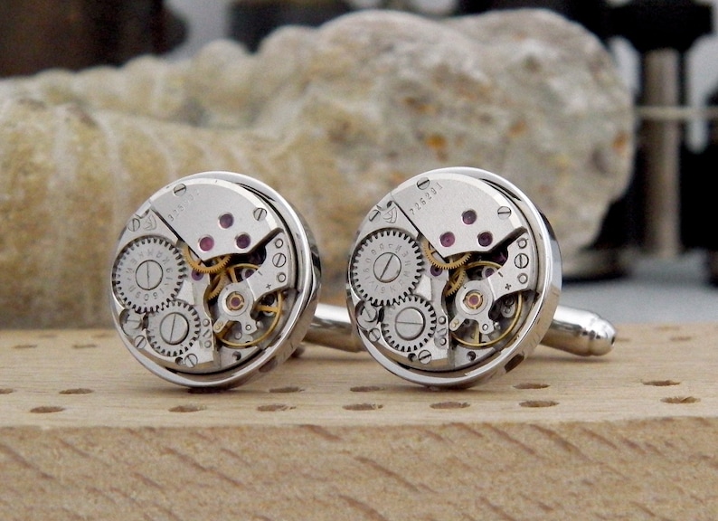 Puede incluir: Un par de gemelos de plata con un dise&ntilde;o circular que presenta engranajes de reloj expuestos y joyas de color rub&iacute;. Los gemelos est&aacute;n sobre una superficie de madera.