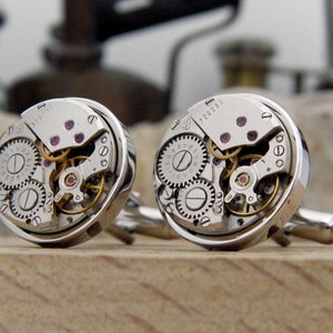 Puede incluir: Un par de gemelos de plata con mecanismos de reloj expuestos. Los gemelos presentan un dise&ntilde;o circular con engranajes visibles y un acabado plateado.
