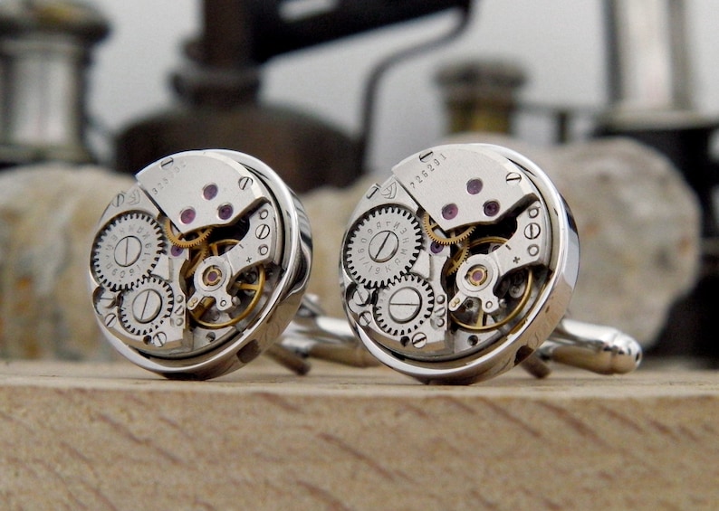 Puede incluir: Un par de gemelos de plata con mecanismos de reloj expuestos. Los gemelos presentan un dise&ntilde;o circular con engranajes visibles y un acabado plateado.