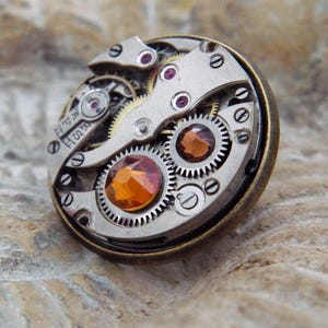 May include: A round, steampunk-style brooch featuring the intricate inner workings of a watch. The brooch has silver-colored gears, ruby-colored jewels, and a large, orange-colored gemstone in the center. The outer rim is a dark bronze color.