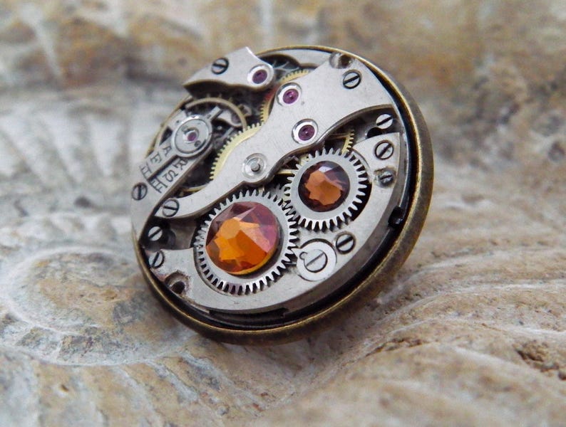 May include: A round, steampunk-style brooch featuring the intricate inner workings of a watch. The brooch has silver-colored gears, ruby-colored jewels, and a large, orange-colored gemstone in the center. The outer rim is a dark bronze color.