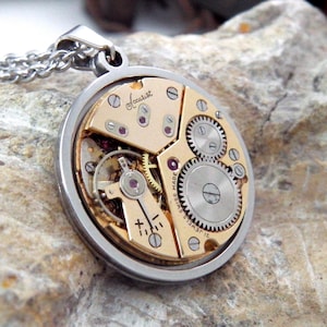 Op de afbeelding: Een zilverkleurige hangerketting met het binnenwerk van een horloge. Het goudkleurige horlogemechanisme met zichtbare tandwielen, schroeven en de merknaam Accurist is te zien. De ketting wordt gepresenteerd op een getextureerd, lichtgekleurd stenen oppervlak.