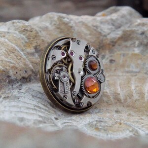 May include: A round, steampunk-style brooch featuring the intricate inner workings of a watch. The brooch has a silver-colored metal base with visible gears, jewels, and an amber-colored gemstone. The outer rim is a bronze color.