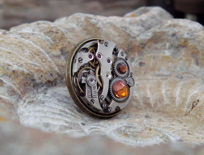 May include: A round, steampunk-style brooch featuring the intricate inner workings of a watch. The brooch has a silver-colored metal base with visible gears, jewels, and an amber-colored gemstone. The outer rim is a bronze color.