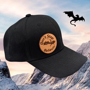 May include: Black baseball cap with a curved brim and a round, tan leather patch. The patch features a dragon silhouette and the words "Don't Stop Believin'". A black dragon silhouette is in the background.