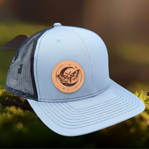 May include: A light blue trucker hat with a black mesh back. The hat features a brown leather patch with a moon and moth design.