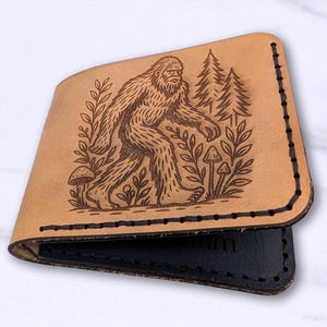 May include: A tan leather wallet with a Bigfoot design. The wallet features a detailed engraving of Bigfoot walking through a forest with trees, mushrooms, and foliage. The interior is black and the edges are stitched.