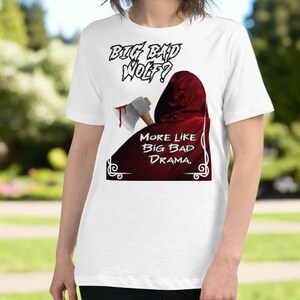 May include: White t-shirt with the text "Big Bad Wolf?" in a gothic font. Below, a red hooded cloak with the text "More Like Big Bad Drama." An axe with blood is also visible.