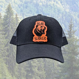 May include: A black baseball cap with a mesh back and a curved brim. Centered on the front panel is an orange patch featuring a black illustration of a roaring bear head and the text BEAR SERKER in a bold, stylized, jagged black font.