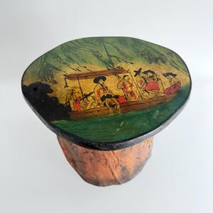 May include: A decorative wooden stool with a round top featuring a painted scene of people in a boat. The top has a black border and rests on a natural, orange-brown base. The scene includes figures in traditional clothing.