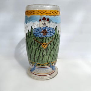 May include: A clear glass drinking vessel with a painted design. The design includes a castle, a bull, and a forest. The vessel has a flared base and a cylindrical body. The text "1700" is visible on the side.