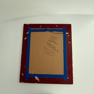 Puede incluir: La parte trasera de una obra de arte enmarcada, con un marco rojo y cinta azul. La obra est&aacute; montada sobre cart&oacute;n, con texto manuscrito. Se adjunta un alambre para colgar la obra.