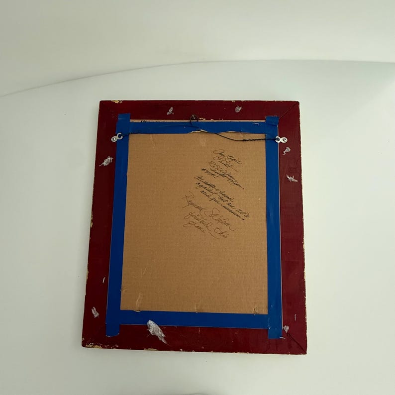 Puede incluir: La parte trasera de una obra de arte enmarcada, con un marco rojo y cinta azul. La obra est&aacute; montada sobre cart&oacute;n, con texto manuscrito. Se adjunta un alambre para colgar la obra.