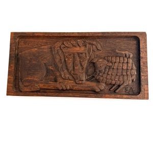 May include: A rectangular dark brown wooden wall plaque featuring a relief carving of a lion lying next to a sheep. The wood grain is visible throughout the piece, and the initials EA are carved into the bottom right corner of the panel.