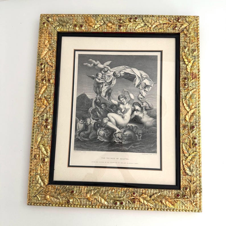 Puede incluir: Una impresi&oacute;n en blanco y negro enmarcada de una escena cl&aacute;sica, con figuras en un paisaje marino. El marco dorado ornamentado tiene un dise&ntilde;o detallado de hojas y bayas con acentos rojos. La impresi&oacute;n se titula "El Triunfo de Galatea."
