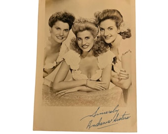 Puede incluir: Una fotografía vintage en tono sepia que muestra a tres personas sonrientes con peinados elaborados, vistiendo blusas claras con mangas de volantes. En la parte inferior de la imagen aparece el texto manuscrito Sincerely Andrews Sisters.