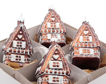May include: A set of four brown glass house ornaments with white glitter detailing, each featuring a peaked roof, windows, and a heart motif. The ornaments are arranged in a partitioned white box with brown paper filler for protection.