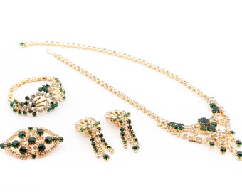 May include: A gold-toned jewelry set featuring a necklace, bracelet, earrings, and a brooch. The pieces are adorned with clear and emerald green rhinestones. The necklace and earrings have a dangling design.