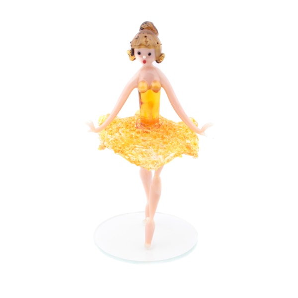 Può includere: Statuina in vetro di una ballerina in posa di danza. La ballerina ha un corpetto giallo e un tutù abbinato. I suoi capelli sono raccolti in uno chignon e si trova su una base circolare trasparente. La statuina è alta circa 15 cm.