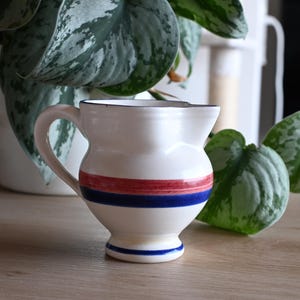 May include: A white ceramic pitcher with a rounded body, a curved handle, and a flared rim. The exterior features horizontal painted stripes in red and blue around the center, with a single blue stripe accenting the base of the pedestal foot.