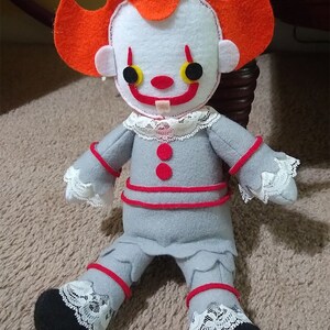 May include: A handmade plush toy of Pennywise the Dancing Clown from the movie "IT". The toy is made of felt and has a red and white costume with a black and white lace trim. The toy has a toothy grin and large, black eyes.