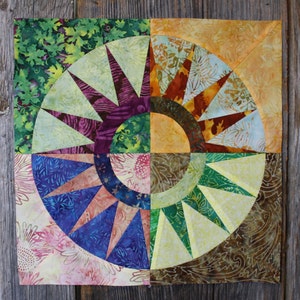 May include: A colourful quilt block with a circular design featuring a starburst pattern in shades of green, blue, purple, and brown. The block is made of fabric with a floral print.