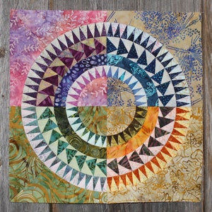 Puede incluir: Un bloque de edredón cuadrado con un diseño circular. El diseño presenta arcos concéntricos en tonos de rosa, morado, azul, verde y naranja, con detalles triangulares. La tela de fondo tiene un patrón de estilo batik.