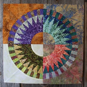 Può includere: Un blocco di trapunta colorato con un design circolare. Il blocco è composto da diversi tessuti con motivi e colori diversi, tra cui viola, verde, blu e marrone. Il centro del blocco è un cerchio bianco.
